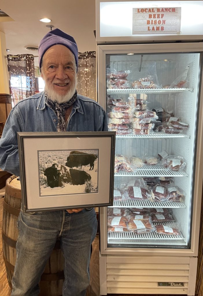 Man with beard holding a picture of buffalo standing next to a cooler with local ranch beef bison and lamb meat in it.