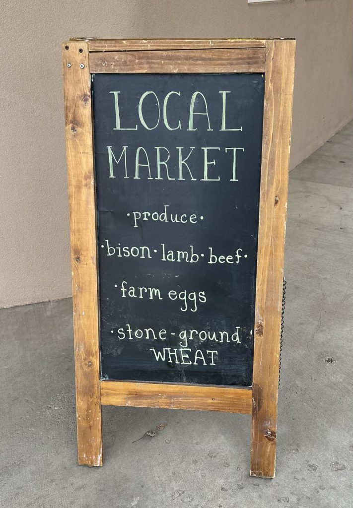 Sign in front of a wall that says Local Market, produce, bison, lamb, beef, farm eggs and stone-ground wheat