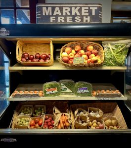 Fruit in a cooler on shelves with onions, apples, eggs, sprouts, tomatoes, carrots, garlic and potatoes and a sign above that says Market Fresh