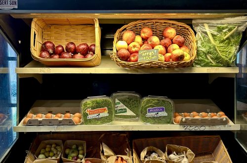 Fruit in a cooler on shelves with onions, apples, eggs, sprouts, tomatoes, carrots, garlic and potatoes and a sign above that says Market Fresh