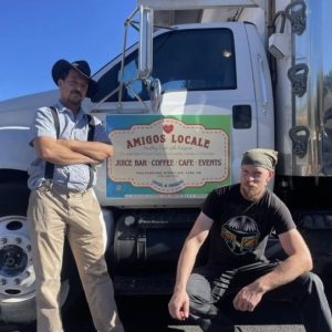 Two men standing next to a refrigerator truck with a sign on the door of the truck that says Amigos Locale, Juice Bar, Coffee, Cafe, Events